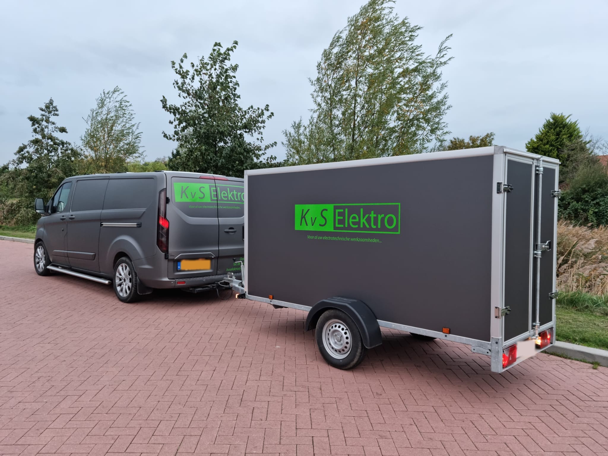 De grijze bedrijfsbus met aanhanger van KvS Elektro, met groene belettering
